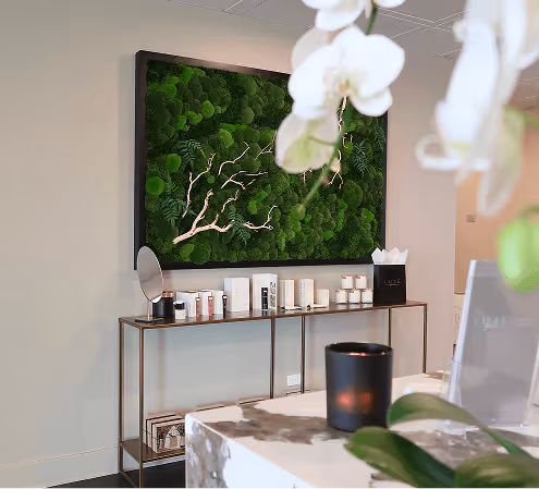 Modern interior with a framed green moss wall art, a console table holding skincare products, and a marble countertop with orchid flowers in the foreground.