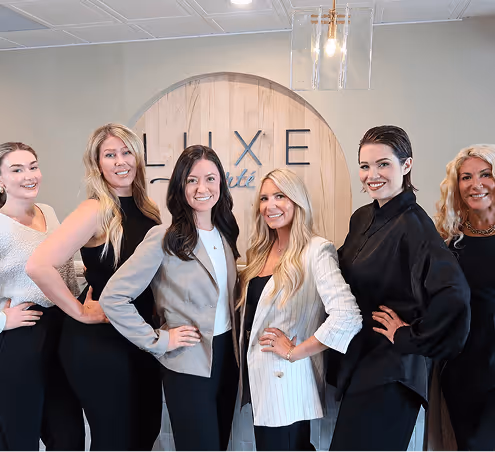 Six women standing side by side, smiling, posing confidently in front of a wooden sign with the text 'Luxe'.