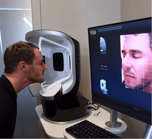Man undergoing a facial skin analysis using a VISIA imaging system with his face positioned inside the scanner and his facial images displayed on a nearby monitor.